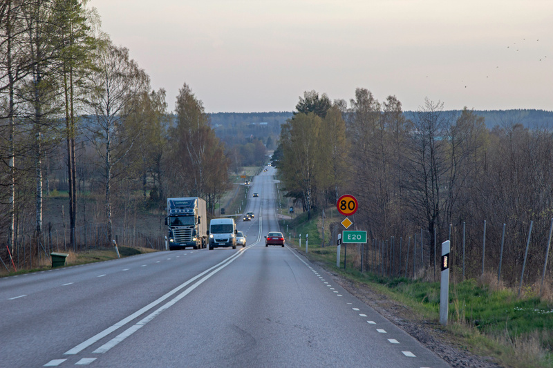Bilden visar en rak landsväg som sträcker sig framåt med fordon som kör i båda riktningarna. På höger sida av vägen syns en hastighetsskylt med fartbegränsningen 80 km/h och en grön vägskylt med texten 'E20'. Längs vägen finns träd och annan vegetati