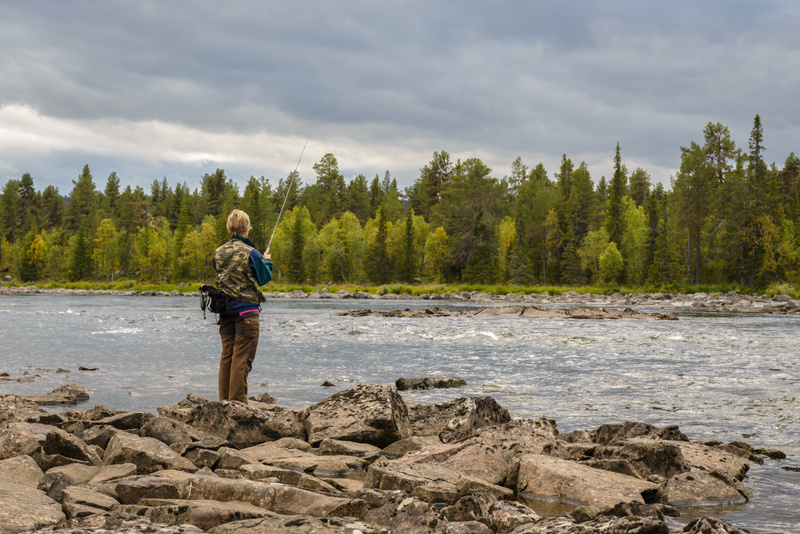 En person står med ryggen mot kameran och fiskar vid kanten av en bred, rinnande flod. Personen bär en kamouflagemönstrad väst över en blå tröja och bruna byxor, och håller ett fiskespö utsträckt över vattnet. Marken framför personen består av stora,