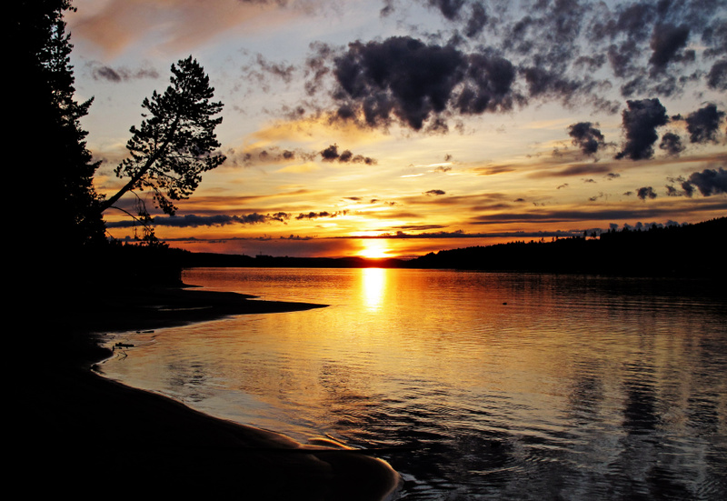 Bilden visar en solnedgång över ett lugnt vattenområde. Solen, som reflekteras starkt i vattnet och skapar ett gyllene sken, är placerad något till vänster om mitten. Horisonten är fylld med varma färger som övergår från gult och orange nära solen ti