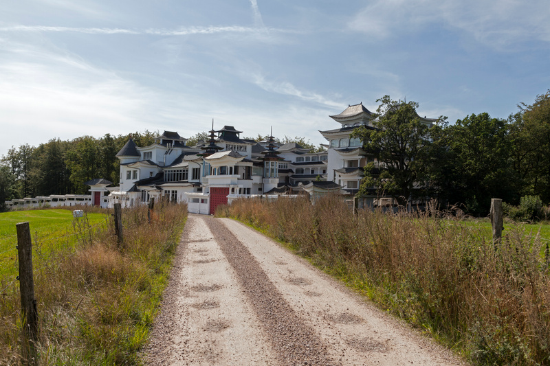 Bilden visar ett stort, vitfärgat hus med arkitektur i asiatisk stil, inklusive svarta, böjda tak och detaljer som pagodliknande torn. Huset är omgivet av grönska, med en gräsmatta framför och täta träd på båda sidor och bakom byggnaden. En grusväg l