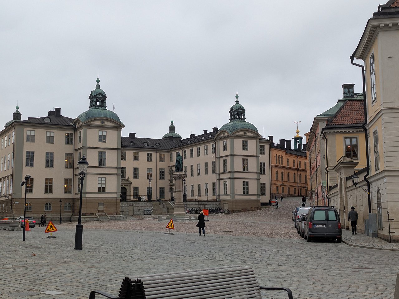 En bild som visar en öppen stenlagd plats omgiven av historiska byggnader. I centrum av bilden står Kungliga Myntkabinettet, en stor ljusgul byggnad med gröna kupoler. Framför byggnaden finns en staty på ett högt fundament. Till vänster om Myntkabine