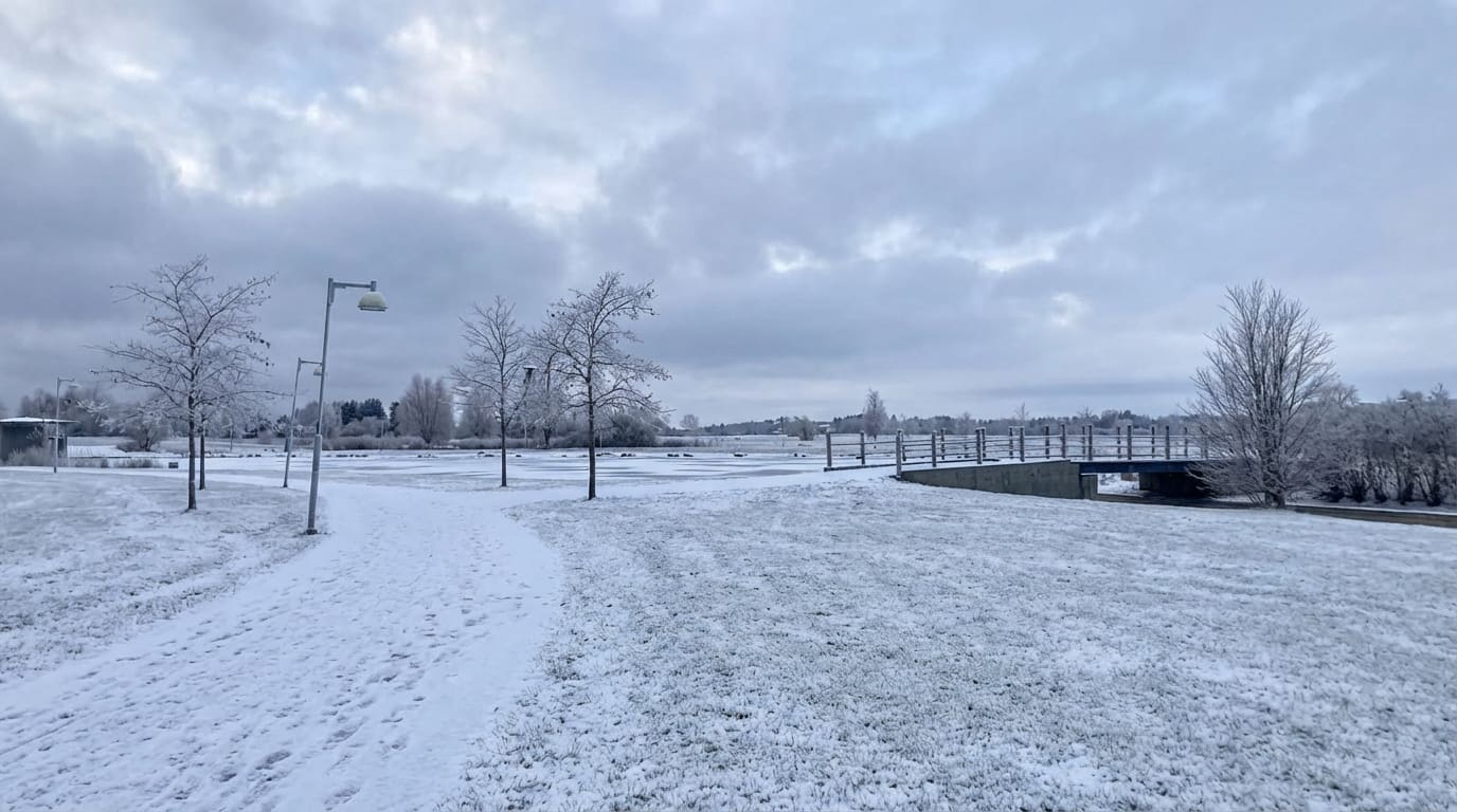 Bilden visar ett snötäckt landskap med en stig i förgrunden som leder mot en sjö eller större vattenyta. Stigen är täckt av ett tunt lager snö och det syns spår i snön. Till vänster om stigen finns flera träd med tunna grenar som också är täckta av s