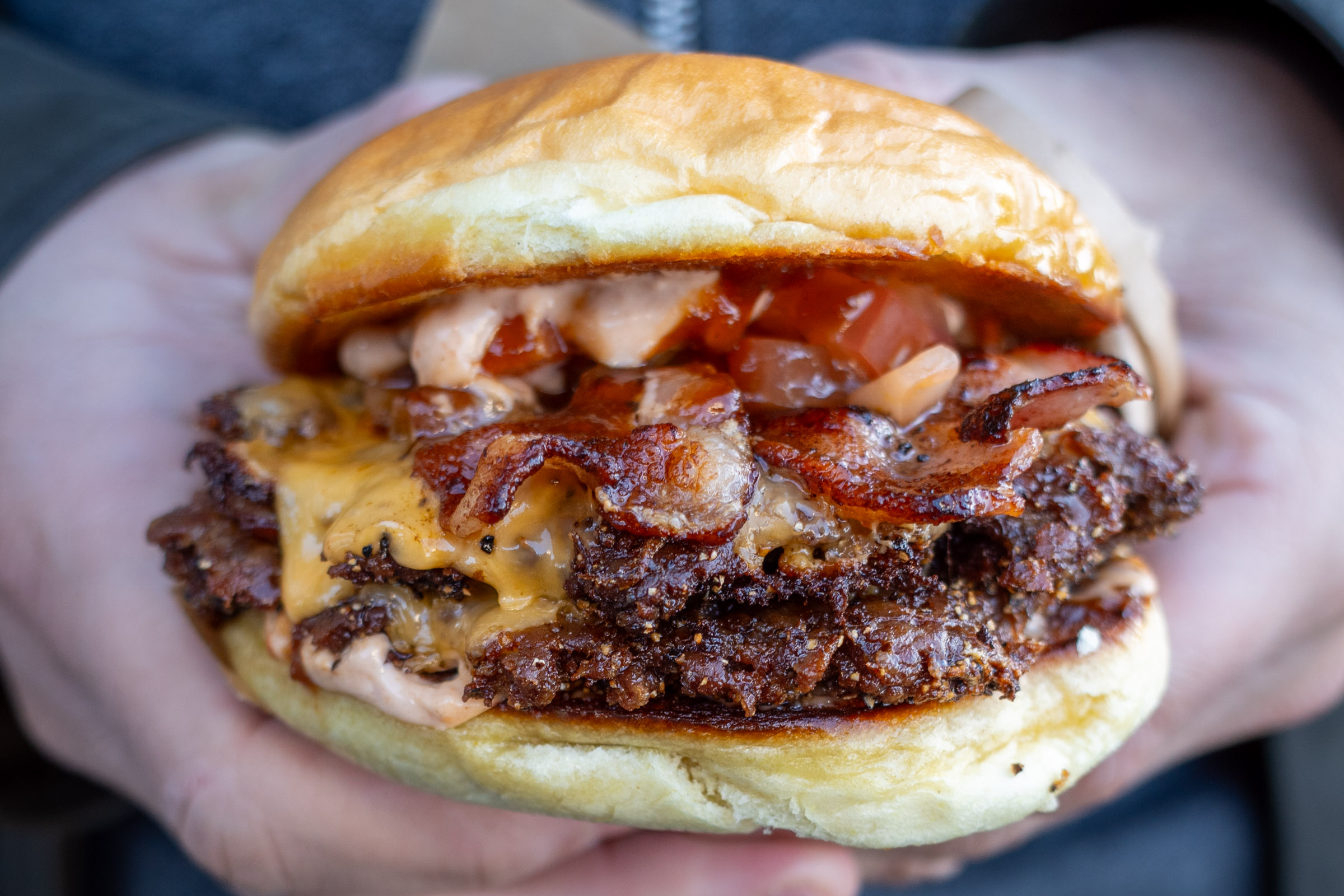 En närbild av en person som håller en stor, fyllig hamburgare med båda händerna. Hamburgaren består av ett rostat hamburgerbröd som omger flera lager. Synliga ingredienser inkluderar två köttbiffar med smält ost, krispiga baconskivor, tärnade tomater