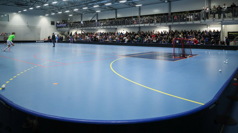 En inomhusbild som visar en stor, ljusblå idrottsplan, troligen för innebandy. En person klädd i grön tröja och vita byxor håller en innebandyklubba och syns på vänster sida av bilden. Planen har vita, orangea, gula och röda linjer samt ett mörkt blå