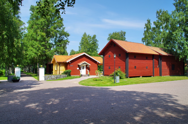 Bilden visar ett landskap med flera byggnader och grönområden under en klarblå himmel. I förgrunden syns en grusväg med skuggor från lövverk. Till vänster finns en allé av lövträd som sträcker sig i bakgrunden, kantad av vita stenpelare med guldfärga