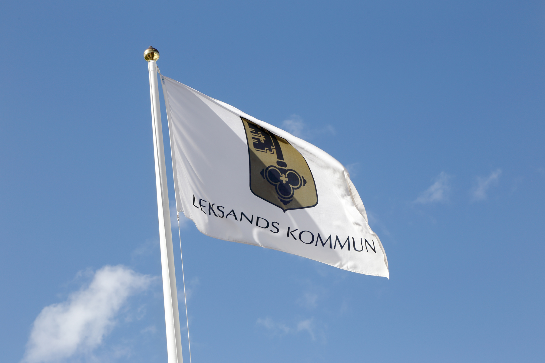 En vit flagga med kommunvapnet för Leksands kommun i brunt, guld och svart, samt texten "LEKSANDS KOMMUN" i svarta versaler, vajar från en vit flaggstång mot en klarblå himmel med några små, vita moln. Flaggan är placerad i den övre halvan av bilden