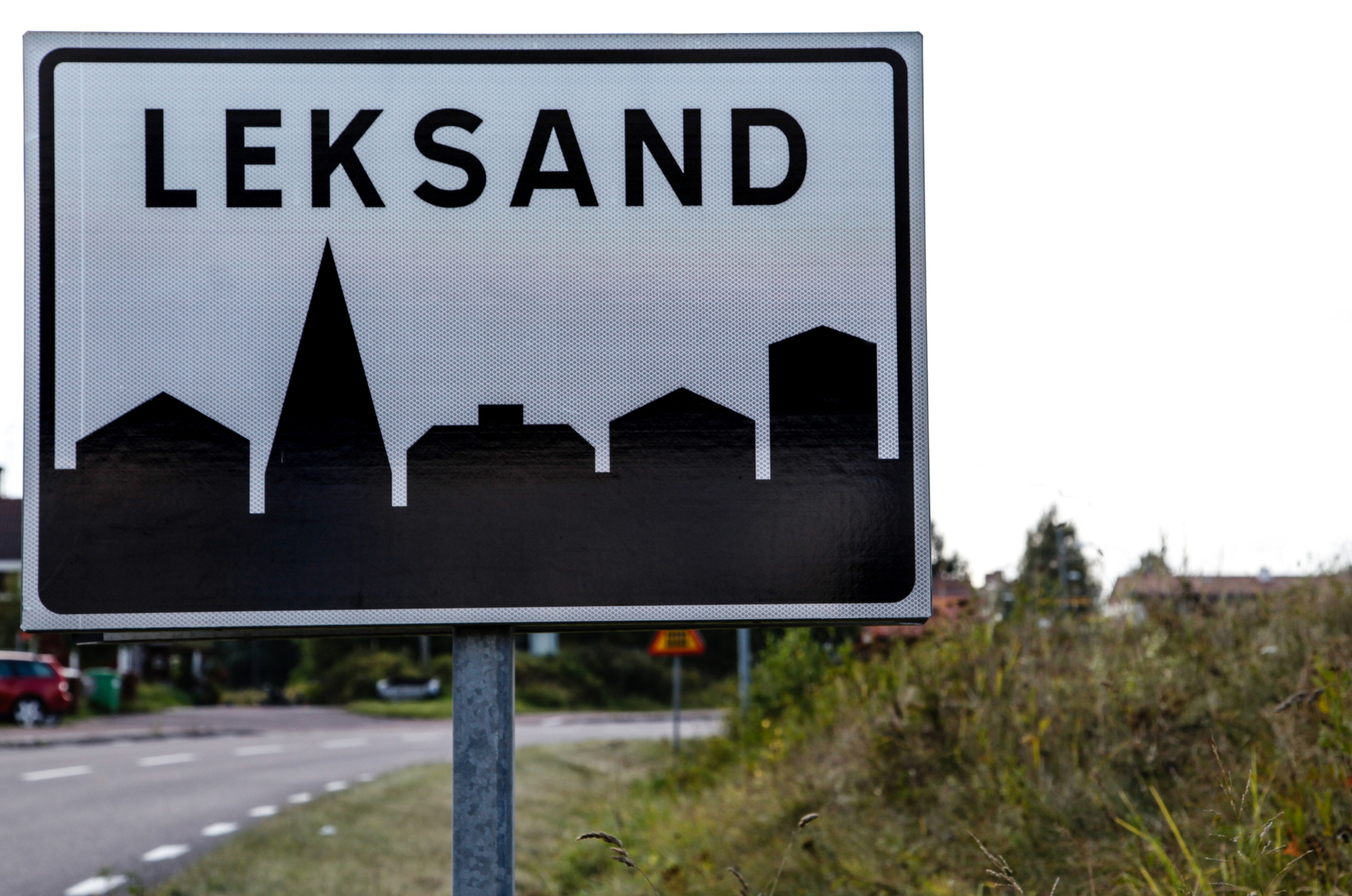 Bilden visar en skylt med texten "LEKSAND" i stora svarta bokstäver på en vit bakgrund. Under texten finns en stiliserad silhuett av byggnader och en kyrkspira i svart. Skylten är monterad på en metallstolpe. I bakgrunden syns en väg med vita vägmark