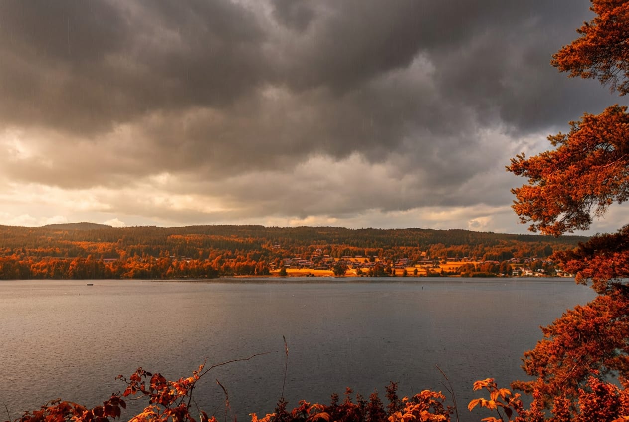 Bilden visar ett vidsträckt landskap med en stor vattenyta i förgrunden och mitten, omgiven av trädklädda kullar och en mindre bebyggelse i bakgrunden. Träden på kullarna och i förgrunden har löv i varma höstfärger, främst orange och rött. En mörk oc