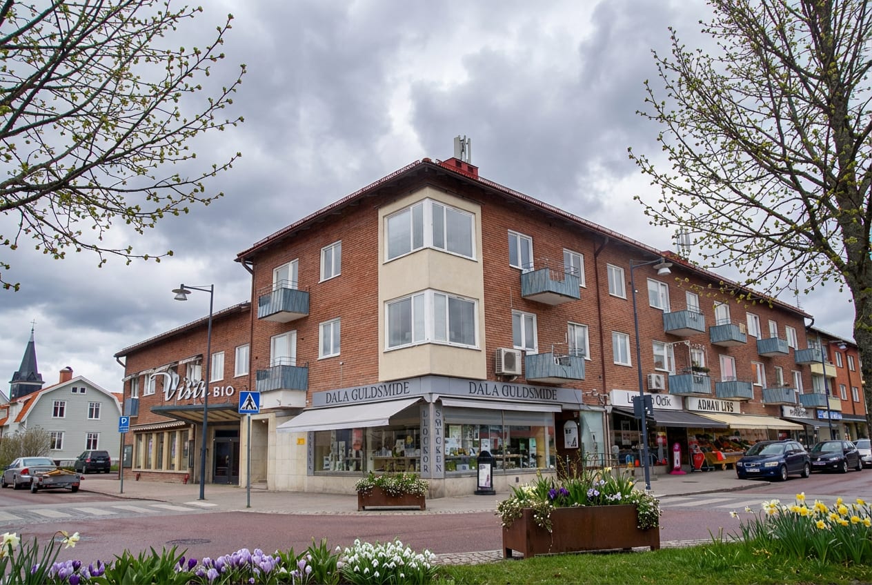 Bilden visar en gatuscen med byggnader, vägar och planteringar under en molnig himmel. Huvudfokus ligger på en röd tegelbyggnad i flera våningar med balkonger. På markplan finns flera butiker, däribland "Dala Guldsmed" och "Adhan Livs". Från vänster