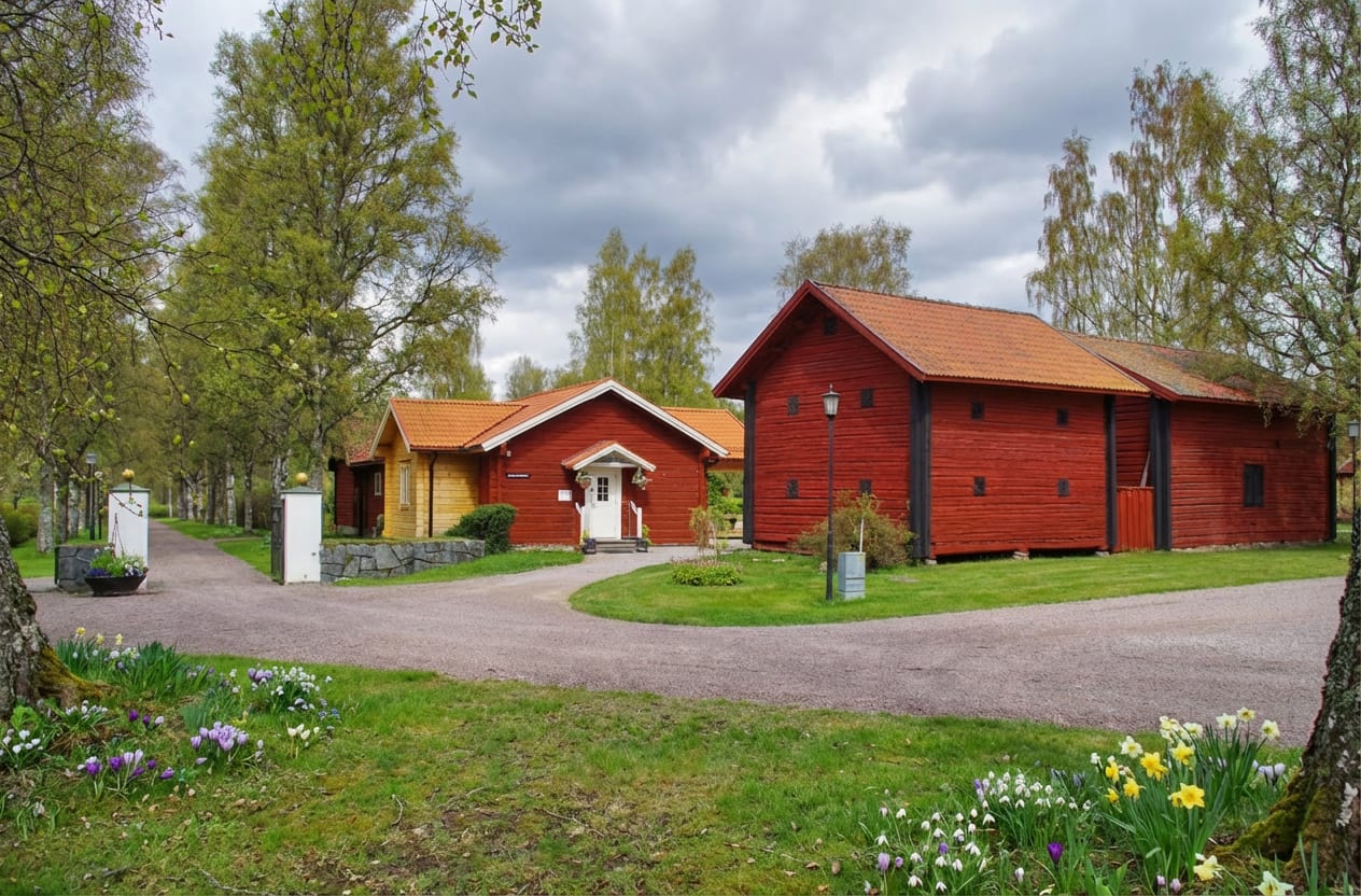 Bilden visar ett landskap med flera byggnader och grönområden under en molnig himmel. I förgrunden syns gräs med vårblommor, inklusive vita snödroppar, lila krokusar och gula påskliljor. En grusväg slingrar sig genom landskapet. På vänster sida av gr