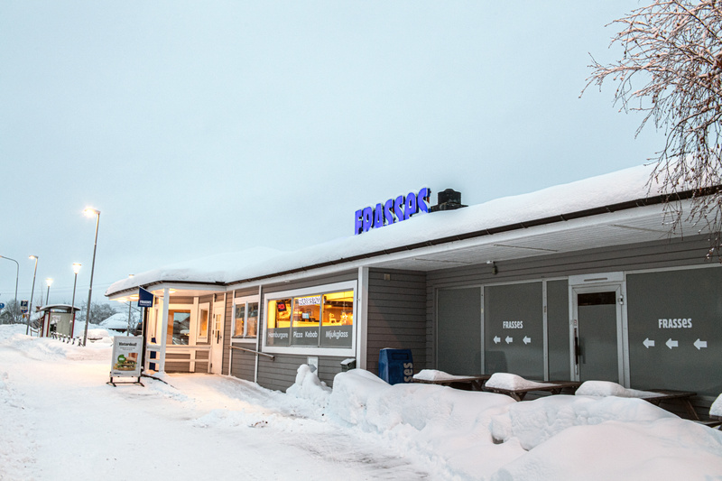 En exteriörbild tagen under dagtid på en snötäckt byggnad som huserar "FRASSES", eventuellt en kiosk eller restaurang, då texten "Hamburgare Pizza Kebab Mjukglass" syns tydligt i ett fönster. Byggnaden är grårandig med vita detaljer och taket är täck