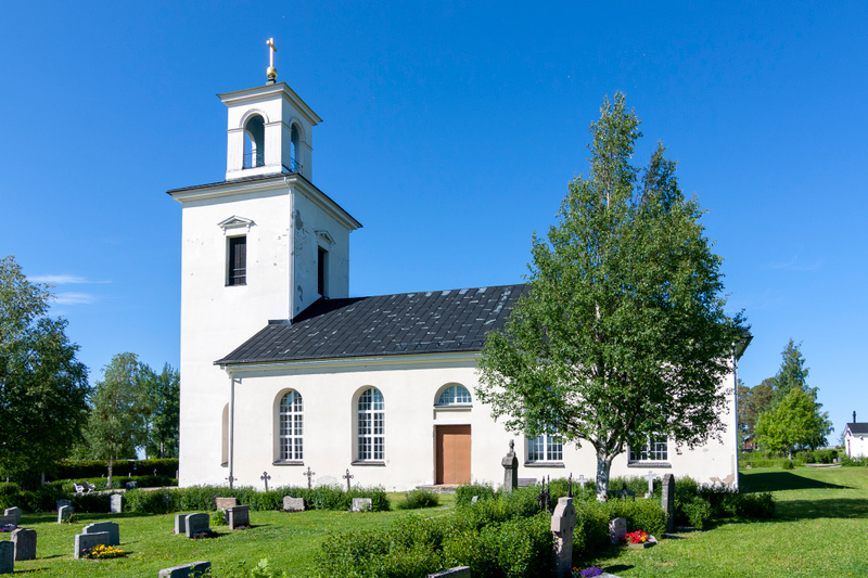 Bilden visar en vit kyrka med en framträdande klockstapel som har ett gyllene kors på toppen. Kyrkan har en mörkgrå takbeläggning och flera välvda fönster med vita karmar. Framför kyrkan syns en kyrkogård med gräsmatta, buskar och flera gravstenar av