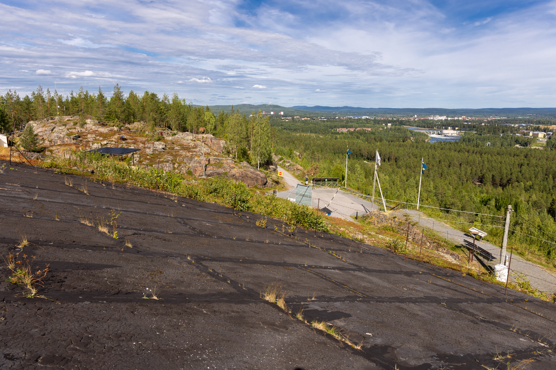 Bilden visar ett landskap taget från en upphöjd position, med en svart, sluttande yta i förgrunden som är täckt med sprickor och sporadiskt växande gräs. I mitten av bilden finns ett bergigt område med träd och buskar, samt en mindre väg som slingrar