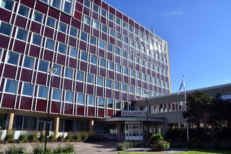 Bilden visar fasaden på en stor byggnad med många fönster. Byggnaden har en mörkröd fasad med vita ramar runt varje fönster. Fönstren är rektangulära och sitter tätt intill varandra i ett rutmönster. Framför byggnaden syns en ingång med en skylt ovan