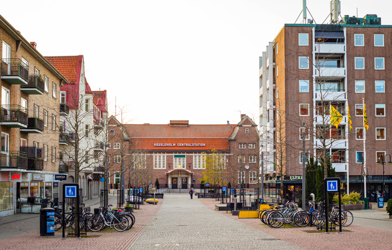 En bild som visar en gågata mellan byggnader med Hässleholms Centralstation i bakgrunden. På vänster sida av gågatan står en rad med flervåningsbostadshus med balkonger, varav en butik med texten "H&M" och en annan med texten "SMYCKA" syns i markplan