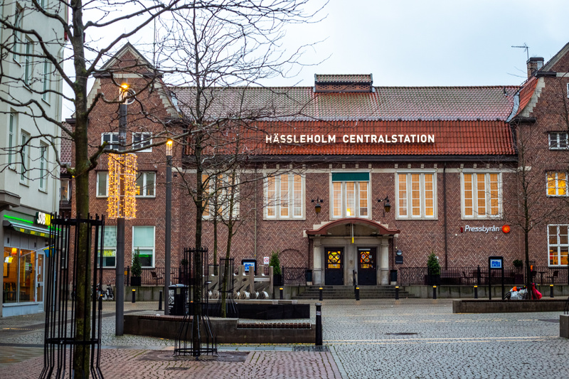 En bild tagen utomhus under dagtid, föreställande Hässleholms Centralstation. Byggnaden har en tegelfasad och rött tak, med 'HÄSSLEHOLM CENTRALSTATION' skrivet i stora vita bokstäver på fasaden. Flera fönster med ljus inifrån är synliga, och framför