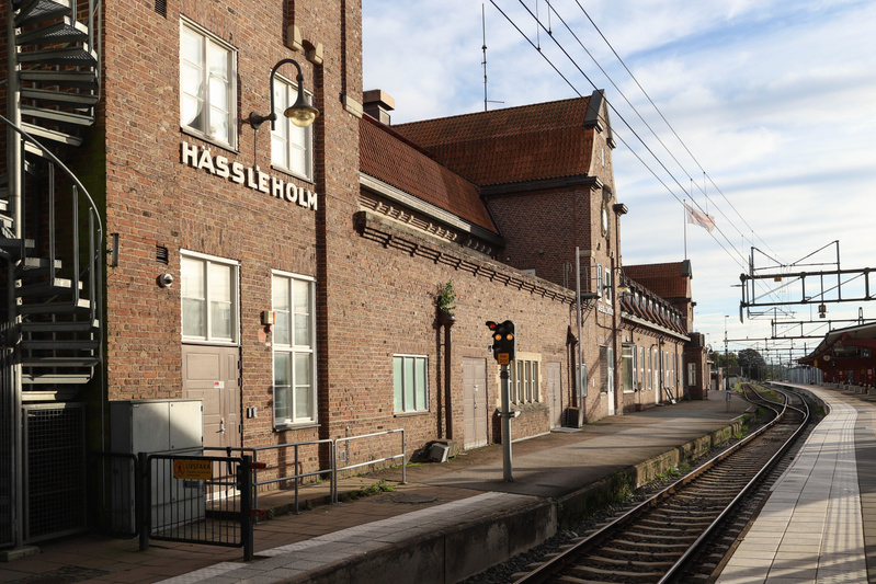 En bild som visar en järnvägsstation med tegelbyggnader, spår och perronger under en delvis molnig himmel. Till vänster syns en stor, röd tegelbyggnad med vita fönsterkarmar och ett brunt tegeltak. På byggnadens fasad står ordet "HÄSSLEHOLM" skrivet