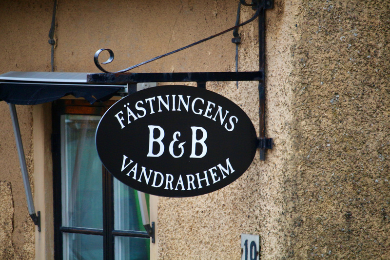 Bilden visar en svart, oval skylt med vit text som fästs på en beigefärgad putsad vägg. Texten på skylten lyder "FÄSTNINGENS B&B VANDRARHEM". Skylten hålls upp av en svartmetallkonstruktion med smidesdetaljer, inklusive en krullad del överst. Till vä