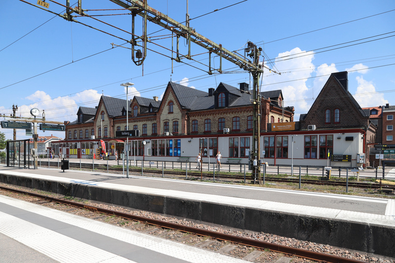 Bilden visar en järnvägsstation från perrongen sett mot stationsbyggnaden. I förgrunden syns räls med vita markeringar på en perrong. Ovanför rälsen och perrongen sträcker sig kontaktledningar för tåg över bilden. Stationsbyggnaden i bakgrunden är te