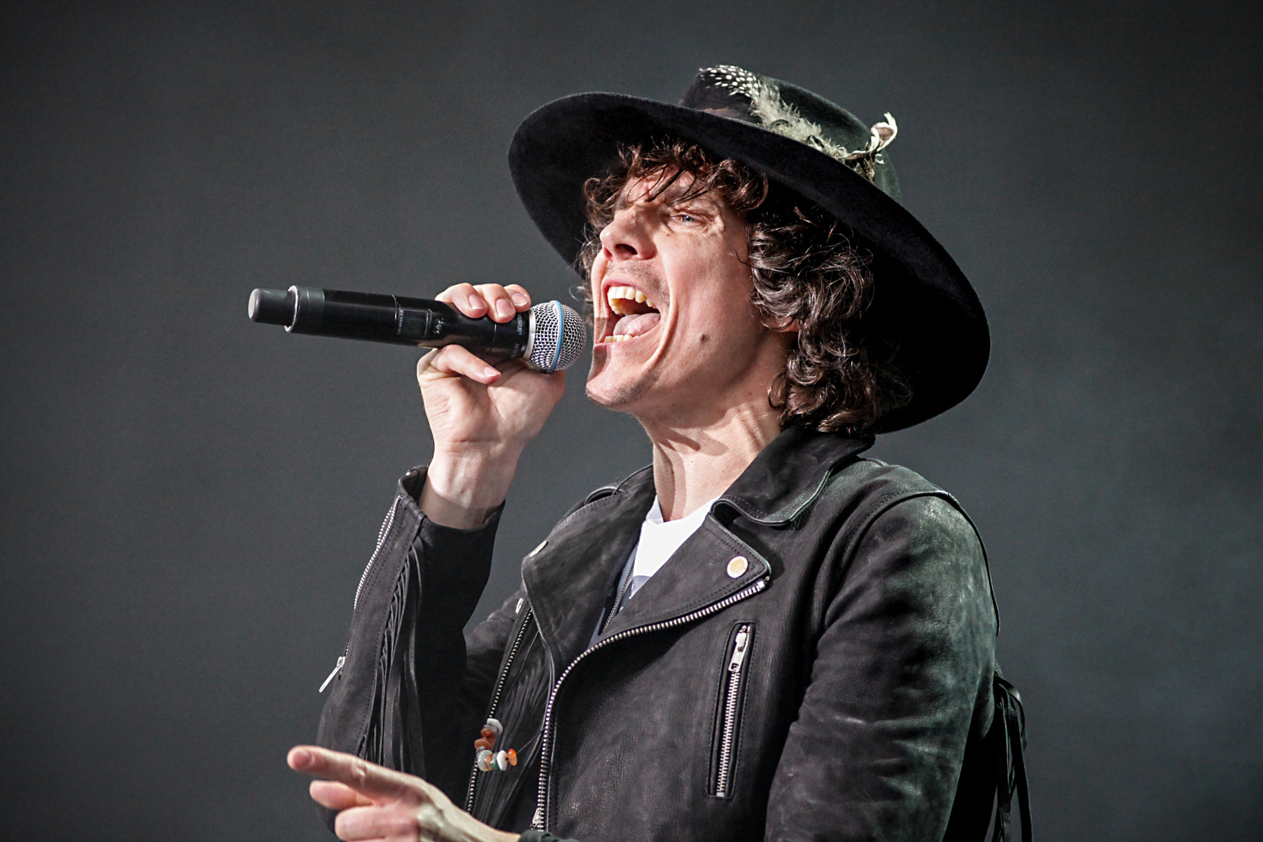 Bilden föreställer en person som sjunger i en mikrofon. Personen bär en svart hatt med fjädrar samt en svart läderliknande jacka med blixtlåsdetaljer och fransar. Under jackan syns en vit t-shirt. Personen har lockigt mörkbrunt hår och ansiktet är vä