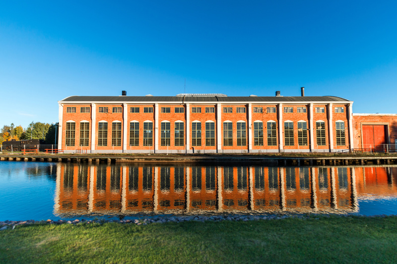En stor, rektangulär byggnad i rött tegel står bredvid en vattenyta. Byggnaden har ett mörkt tak och en lång rad av höga, vertikala fönster med många små rutor längs hela framsidan. Mellan fönstren finns vita vertikala pelare. Vattnet reflekterar byg