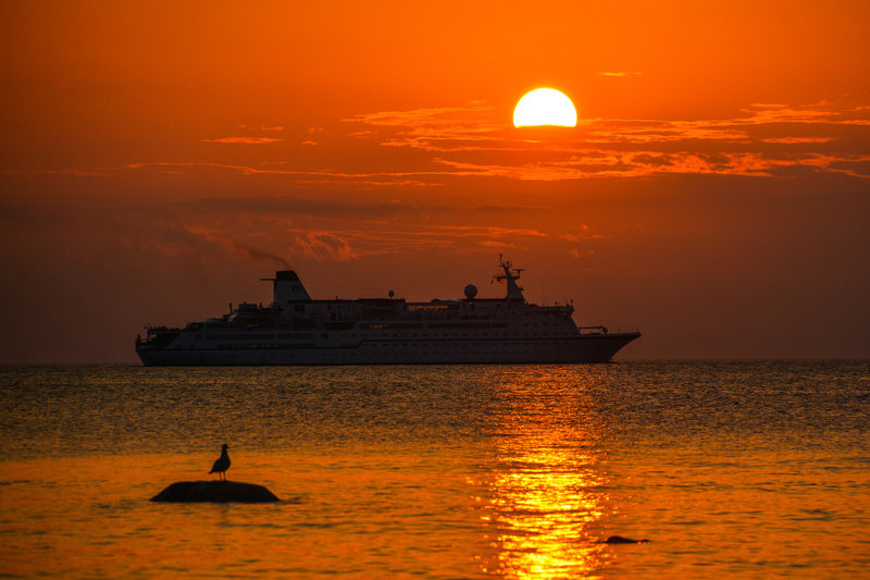 Bilden visar ett stort fartyg, troligtvis ett kryssningsfartyg eller en färja, på öppet vatten under en solnedgång. Solen är synlig som en halv cirkel av klart gult och orange ljus mot en himmel som domineras av orange och mörka moln. Vattenytan refl