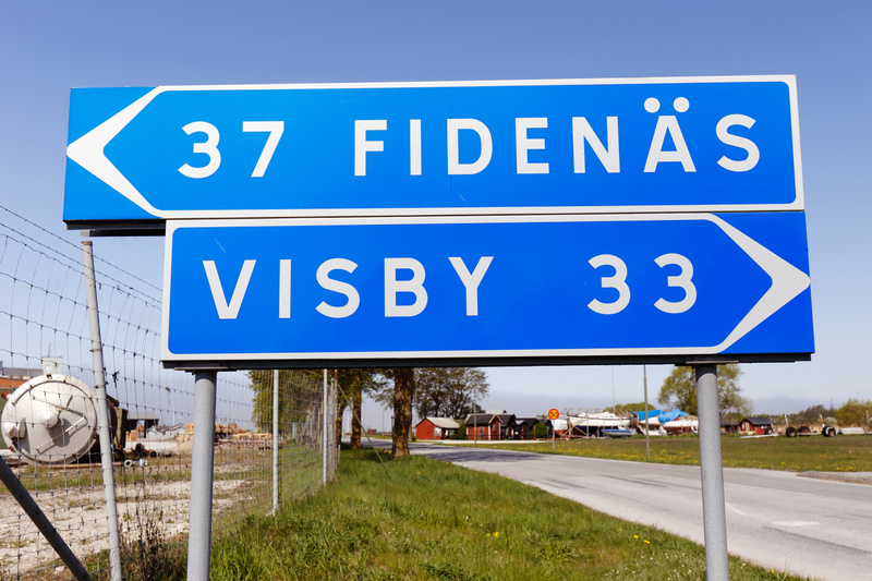 En bild tagen utomhus under dagtid som visar en blå vägskylt monterad på två stolpar. Den övre delen av skylten har texten "37 FIDENÄS" med en vit pil som pekar åt vänster. Den nedre delen av skylten visar "VISBY 33" med en vit pil som pekar åt höger