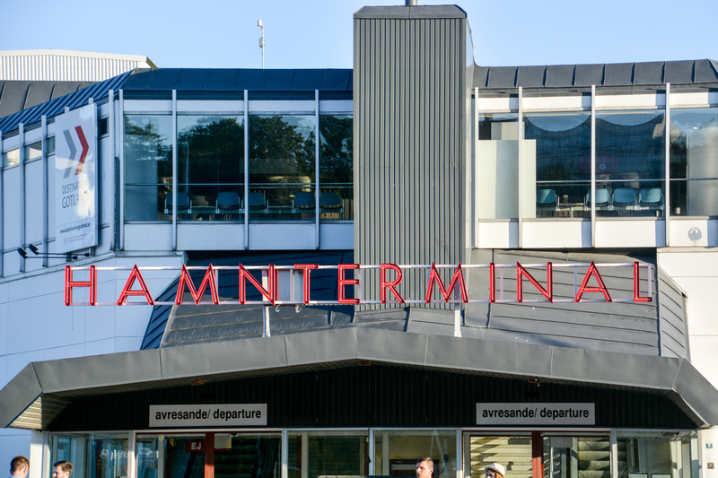 Bilden visar fasaden på en byggnad med texten "HAMNTERMINAL" i röda neonbokstäver centrerad ovanför ingångspartiet. Byggnaden har stora fönsterpartier på övervåningen som reflekterar omgivande träd och himmel. Innanför fönstren syns möbler och stolar