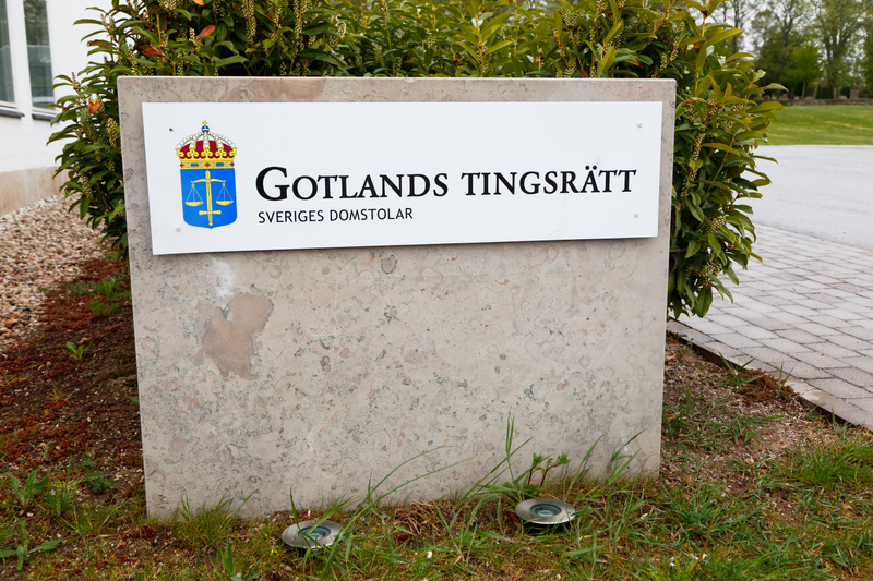 En bild tagen utomhus från en något högre vinkel. Centrerat i bilden står en rektangulär skylt gjord av ett ljust grått, flammigt material som liknar sten. På denna sten sitter en vit rektangulär plåt med texten "GOTLANDS TINGSRÄTT" i stora svarta bo