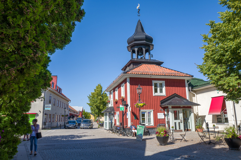 En bild tagen utomhus under dagtid, föreställande ett stadstorg eller en gata under soligt väder. I förgrunden till vänster syns grönska från ett träd. Huvudmotivet i bilden är en röd byggnad med en svart spira och en väderflöjel med formen av en tup