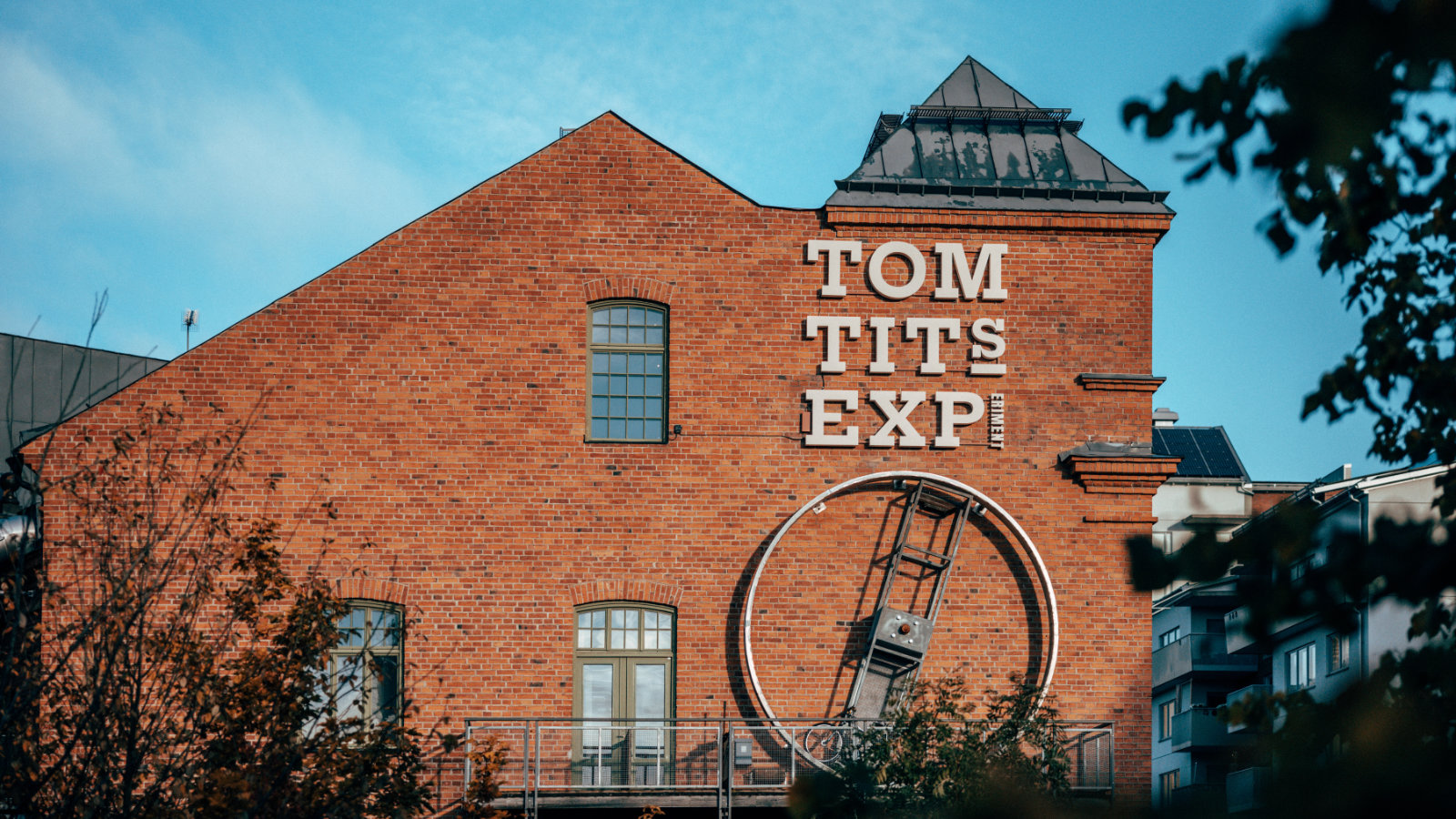 En stor röd tegelbyggnad med vita bokstäver som bildar 'TOM TITS EXPERIMENT' på fasaden. Byggnaden har flera fönster, en balkong med ett grått räcke och en stor mekanisk konstruktion i form av ett hjul fäst vid väggen. Delar av träd med löv är synlig