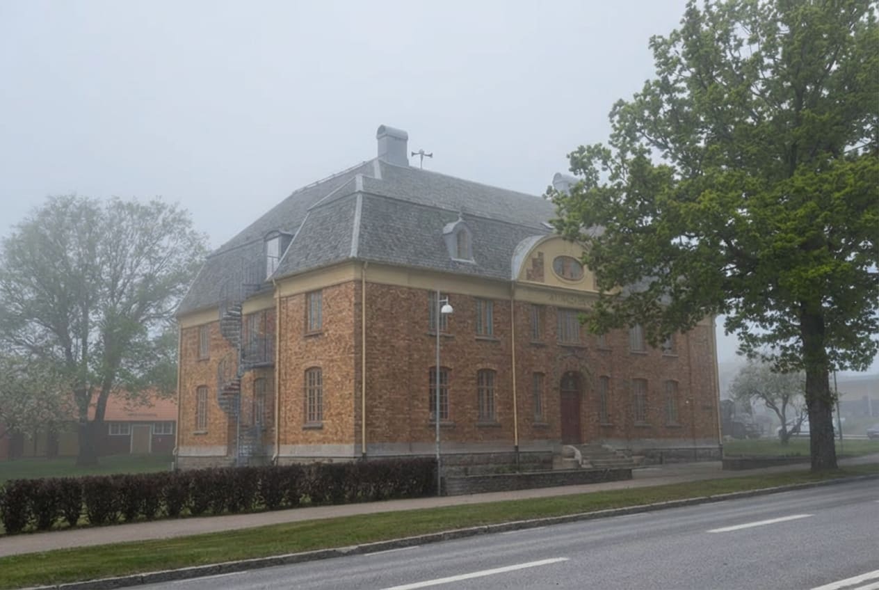 En stor tegelbyggnad med grått tak och en synlig yttertrappa av metall på vänster sida. Byggnaden står intill en väg med gräsmatta och buskar emellan. Tjock dimma ligger över landskapet, och träd med grönskande lövverk syns på båda sidor om byggnaden