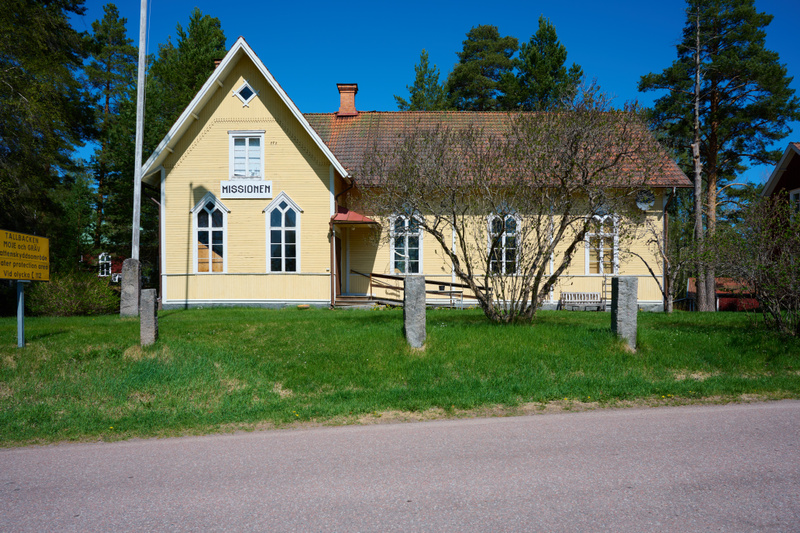 En gul byggnad med skylten "MISSIONEN" placerad på en gräsbevuxen kulle intill en väg. Himlen är klarblå, och barrträd syns i bakgrunden. Ett uthus kan anas till höger om huvudbyggnaden. Skylten till vänster om byggnaden är delvis skymd och varnar fö