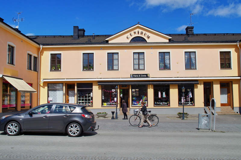 En gata med gatsten framför en byggnad med ljust gulbeige fasad. Byggnaden har butiker ('Hans & Greta') på bottenvåningen. En grå bil är parkerad till vänster, och en person leder en cykel på trottoaren till höger. Himlen är blå med några vita moln.