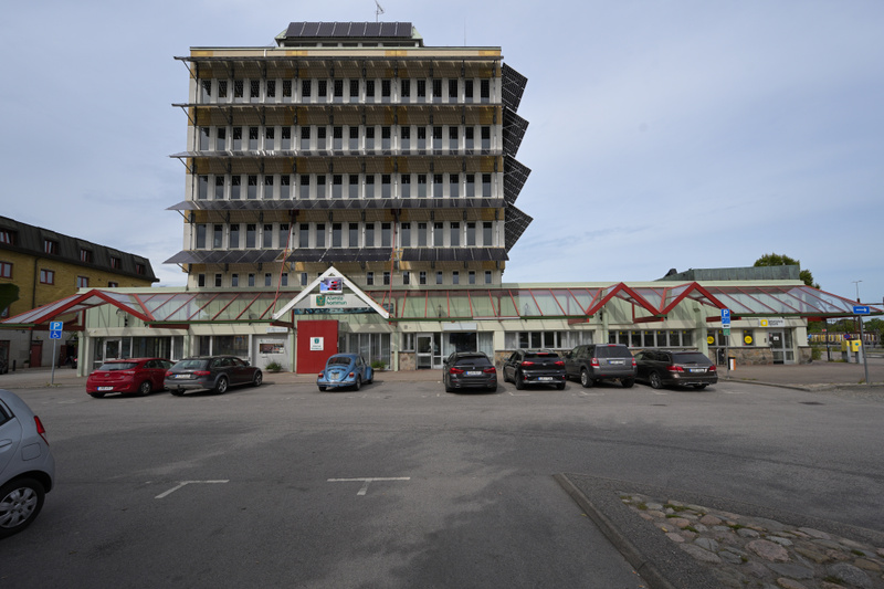 En stor, flervåningsbyggnad med solpaneler på fasaden och taket, dominerar bildens mitt. Framför byggnaden finns en parkeringsplats med flera parkerade bilar. Byggnadens nedre del har en glasinbyggd entré och därbakom ytterligare byggnader skymtar