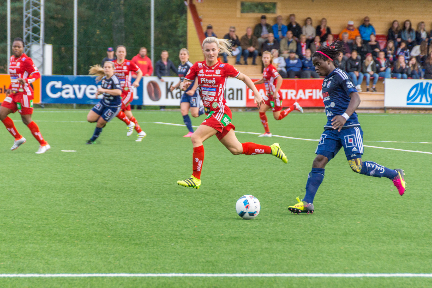 Bilden visar en kvinnlig fotbollsmatch utomhus på en grön fotbollsplan. Flera spelare i röda och blå dräkter springer på planen, och en fotboll syns nära fötterna på en spelare i blått. I bakgrunden sitter åskådare på läktaren, och flera reklamskylta