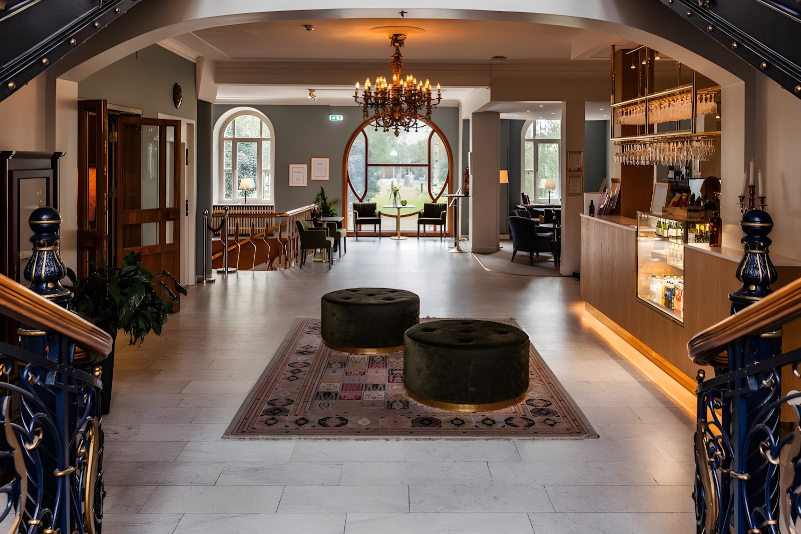 Bilden visar ett inomhusmiljö med en foajé eller hotellobby, sett från nedre delen av en trappa. I förgrunden syns ett mönstrat golv och en matta med två cylinderformade fotpallar i mörk färg. En stor kristallkrona hänger i taket, och framför den fin