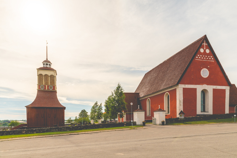 En röd kyrkobyggnad med ett kopparbelagt tak och vita detaljer står bredvid en fristående klockstapel. Framför byggnaderna syns en asfalterad väg och en gräsmatta. Himlen är ljus och molnig.