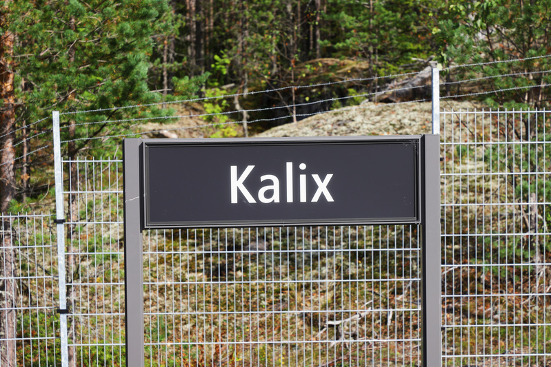 En svart skylt med texten "Kalix" i vitt står framför ett metallstaket med taggtråd. Bakom staketet syns gröna träd och klippor under en solig himmel.