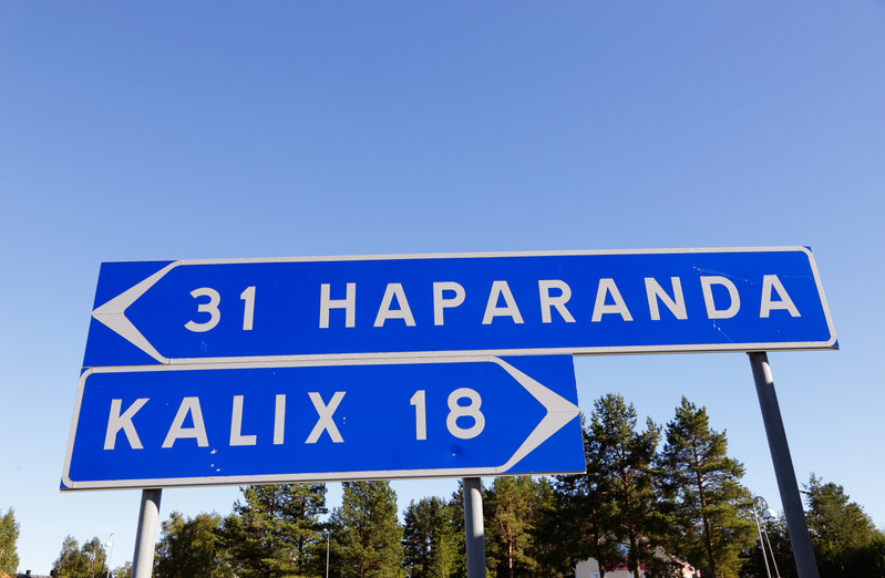 En blå vägskylt med vit text på två rader med texten "31 HAPARANDA" och en pil som pekar åt vänster på övre skylten, och "KALIX 18" med en pil som pekar åt höger på den undre. Skylten står mot en klarblå himmel med tallar i bakgrunden.
