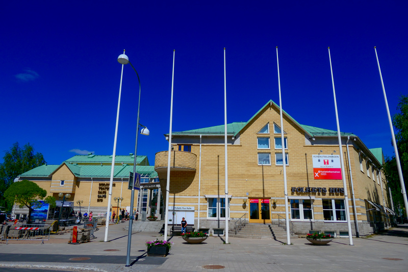 En gul tegelbyggnad med gröna tak och flera fönster är central i bilden. Framför byggnaden syns flaggstänger, gatlyktor och blomsterurnor. Människor passerar på en trottoar framför byggnaden. En skylt med texten "Folkets Hus" är synlig.