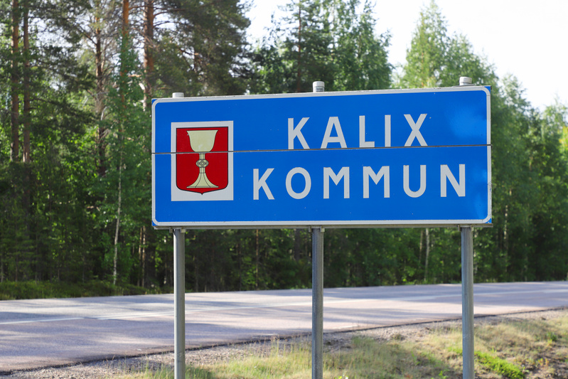 En blå skylt med vit text som läser "KALIX KOMMUN" och Kalix kommunvapen med en kalk på röd botten. Skylten är placerad på två stolpar vid sidan av en asfaltväg. I bakgrunden syns skog med gröna träd och blå himmel.