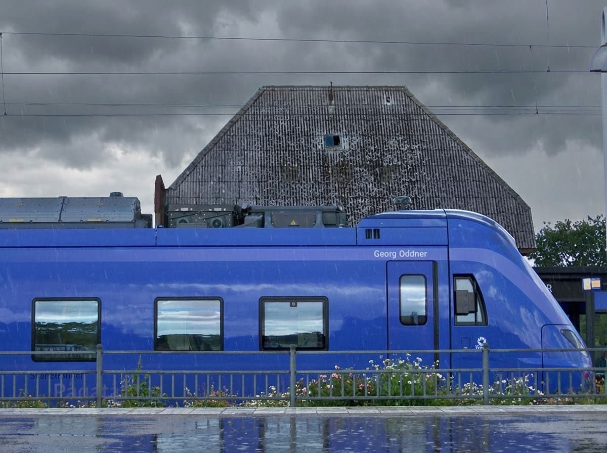 En blå modern tågvagn är delvis synlig vid en plattform under regnigt och molnigt väder. Vagnen reflekterar omgivningen i sina fönster. I bakgrunden syns ett grått hus och en stolpe, samt en blomrabatt längst med plattformskanten.