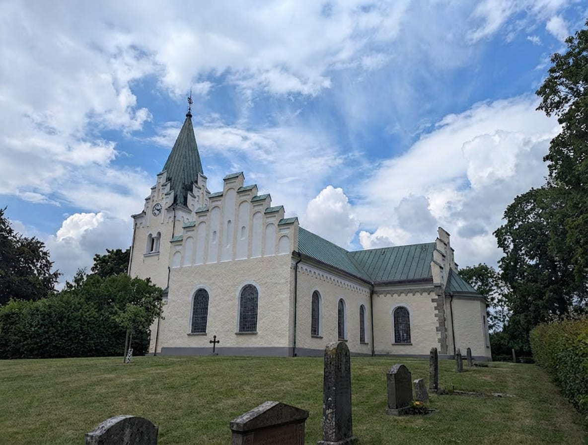 En ljusgräddfärgad kyrka med ett grönt tak och en spetsig tornspira syns från sidan. I förgrunden är det en grön gräsmatta med flera grå gravstenar utspridda. I bakgrunden syns grönska från träd och en blå himmel med vita moln.