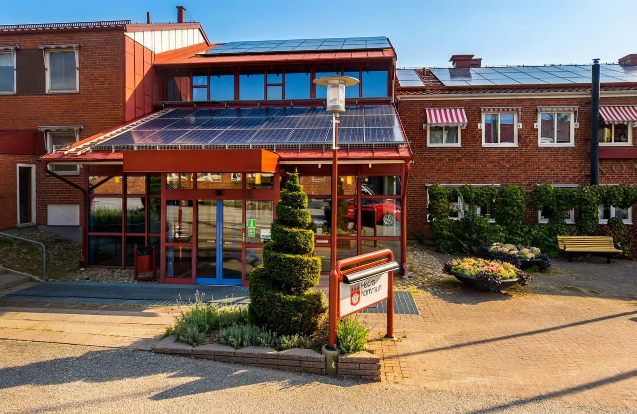 En röd tegelbyggnad med stora glaspartier och solpaneler på taket. Framför byggnaden finns gångplattor, planteringar, en formklippt buske, en gatlampa och en skylt med texten "Hörby kommun". Solen skiner och himlen är klarblå.
