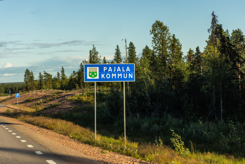 En blå skylt med texten ”PAJALA KOMMUN” och kommunvapnet framför en skogsklädd backe vid sidan av en asfalterad väg med vit mittlinje. En mindre blå parkeringsskylt syns även i bakgrunden.