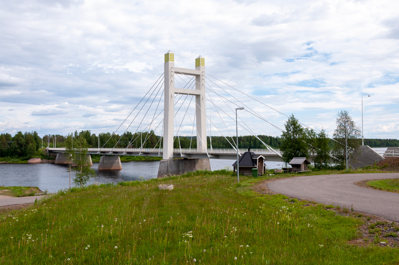 En bro sträcker sig över ett vattendrag med trädklädda stränder under en mulen himmel. I förgrunden syns en gräsbevuxen sluttning med en asfalterad väg som svänger åt höger, samt några små byggnader nära vattnet till höger om bron.