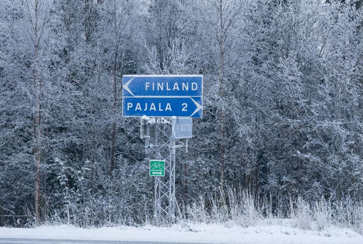 En vägskylt med orden "FINLAND" och "PAJALA 2" syns, täckt av snö. Skylten är installerad på en metallstolpe och bakom den finns ett snötäckt skogslandskap. Vägen i förgrunden är även den snötäckt.
