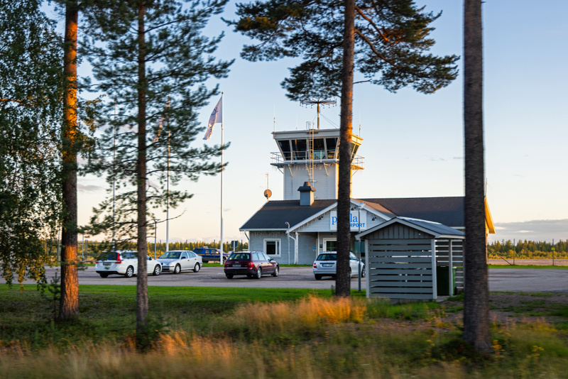 En bild av en flygplatsbyggnad med ett kontrolltorn, omgiven av träd och parkerade bilar. Ett flertal flaggstänger med flaggor syns bredvid byggnaden. Marken består av gräs och grus. Himlen är ljus med några moln.