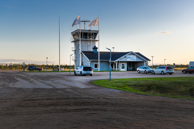 En bild utomhus som visar Pajala flygplats. En kontrolltorn byggnad i grått och vitt med en intilliggande byggnad med texten "pajala airport" på fasaden. Ett flertal bilar är parkerade utanför och två flaggor vajar på en stång vid kontrolltornet. Mar